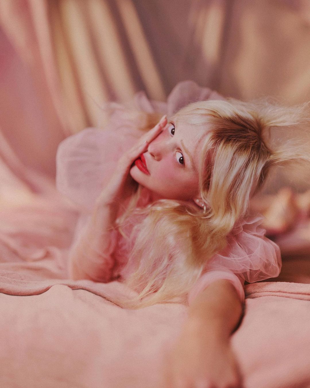 A young adult woman with pale skin and platinum blonde hair poses in a pink ruffled dress and pink lipstick in a soft-focus portrait.