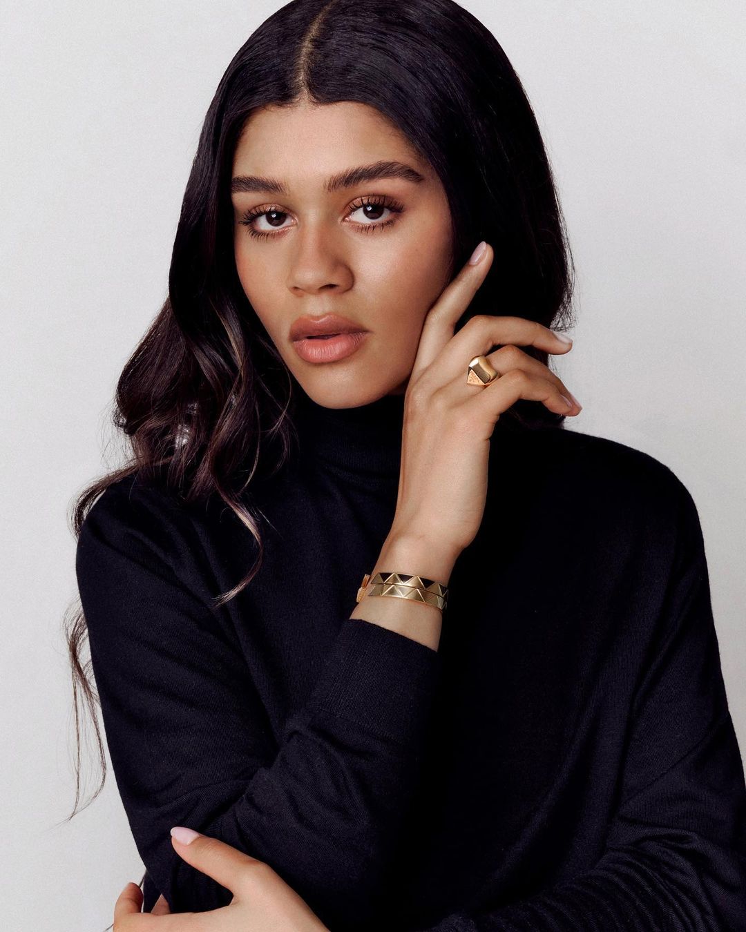 Close-up portrait of a young adult woman with dark hair in a black turtleneck sweater, wearing gold jewelry against a white background.