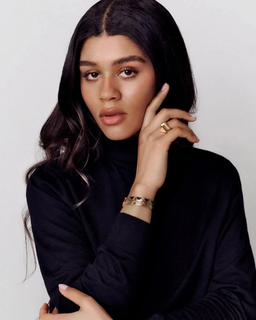 Close-up portrait of a young adult woman with dark hair in a black turtleneck sweater, wearing gold jewelry against a white background.