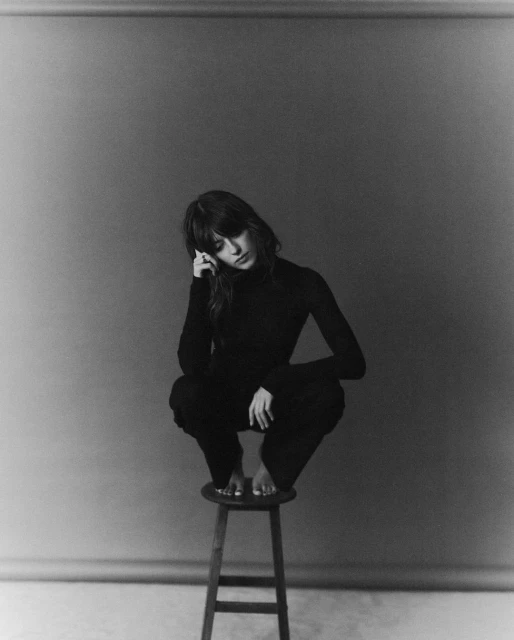 Black and white studio portrait of a woman in a dark turtleneck and pants, squatting on a wooden stool with a neutral background.