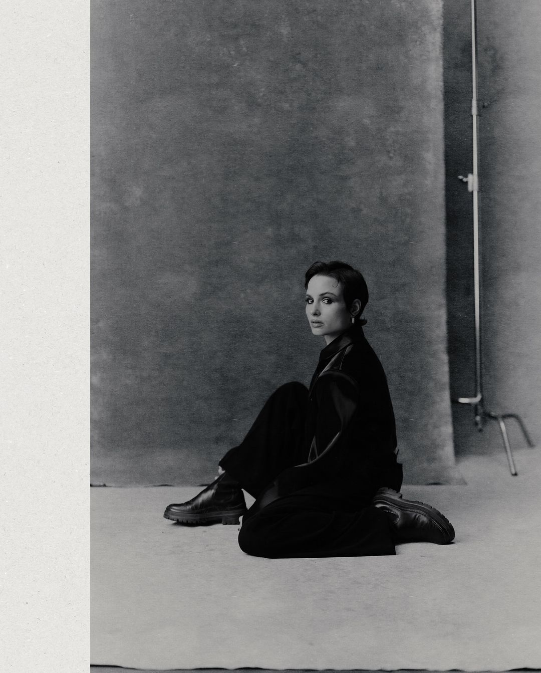 A woman in a black outfit poses seated in front of a mottled grey backdrop in a studio shot in black and white with a vertical composition.