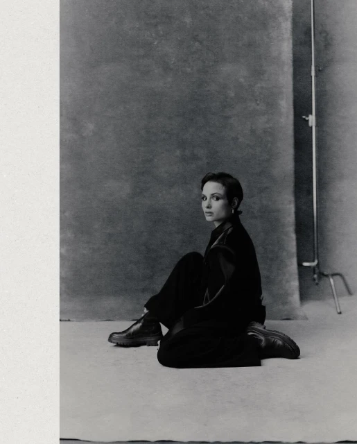 A woman in a black outfit poses seated in front of a mottled grey backdrop in a studio shot in black and white with a vertical composition.