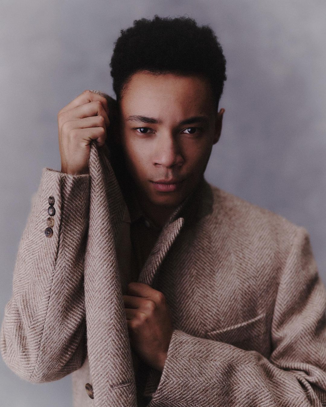 Portrait of a curly-haired adult male in a herringbone blazer, looking at the camera with a serious expression, against a grey backdrop.