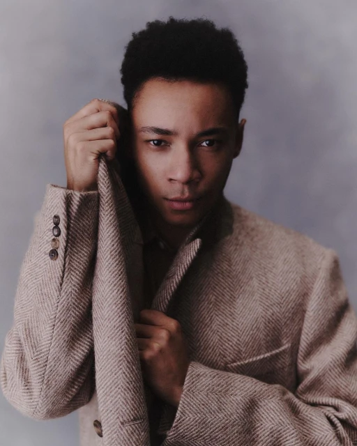 Portrait of a curly-haired adult male in a herringbone blazer, looking at the camera with a serious expression, against a grey backdrop.