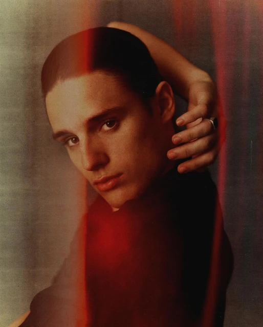 Portrait of a young adult man with short hair, looking at the camera, hand on head, against a textured background with red accents.