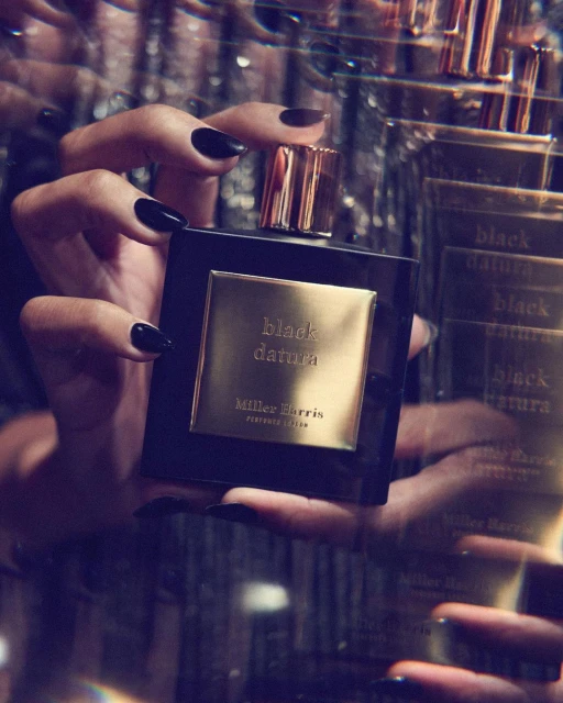 Hands holding a black perfume bottle with a gold label that reads 'black datura Miller Harris' against a dark, sparkly background.