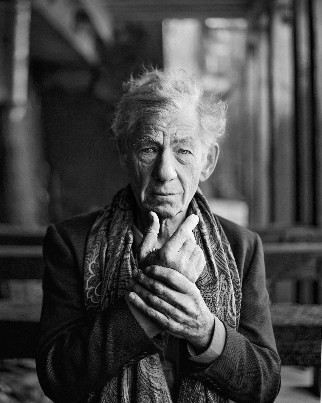 Black and white portrait of an elderly man wearing a patterned scarf and jacket, posed in front of a dark, wood-paneled background.