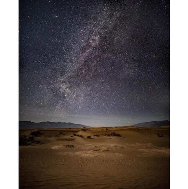 Expansive nighttime desert landscape featuring rolling sand dunes under a starry sky, with the Milky Way visible overhead.