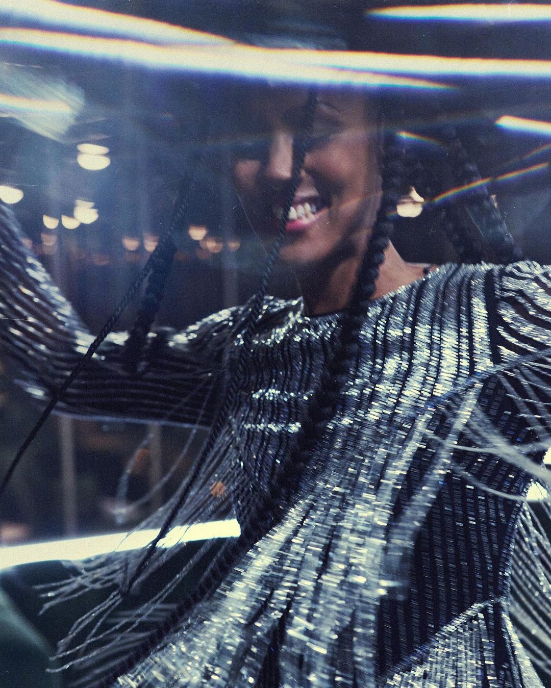 Smiling woman with dark hair in braids wearing a shiny, textured top, seen through a blurred, refractive lens.