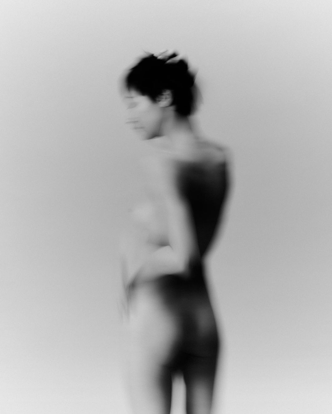 Monochrome portrait of an out-of-focus adult woman with short dark hair looking right, photographed indoors against a light backdrop.