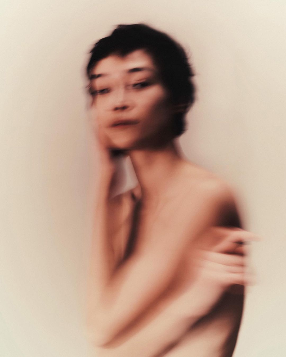 Soft focus portrait of a woman with short dark hair and arms crossed, set against a soft cream background. Intimate and blurred.