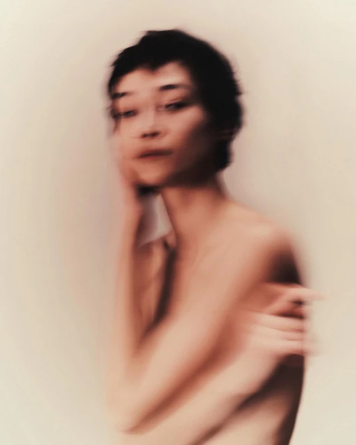 Soft focus portrait of a woman with short dark hair and arms crossed, set against a soft cream background. Intimate and blurred.