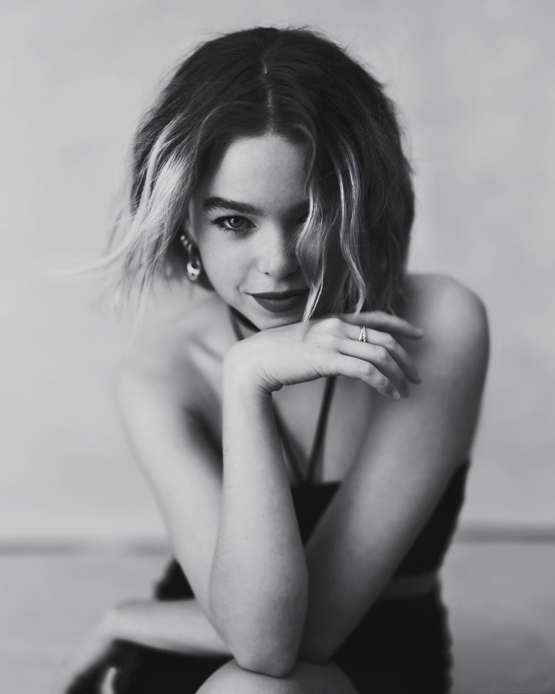 Black and white portrait of a young woman with wavy bob haircut, wearing a crop top and jewelry, posing with hands near her face.