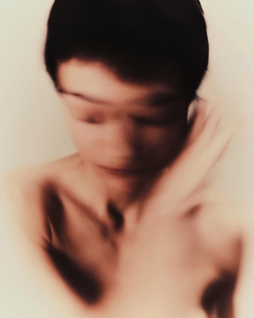 Blurred portrait of a person with short dark hair against a light background, exhibiting soft focus and limited color palette.