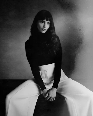 Portrait of a woman with dark hair wearing a black turtleneck and high waisted white trousers against a textured background.