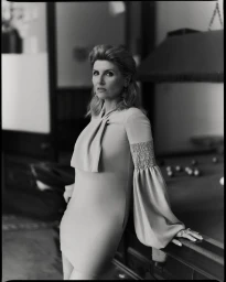 Monochrome portrait of a woman leaning against a pool table wearing a long sleeve dress with smocked cuffs.