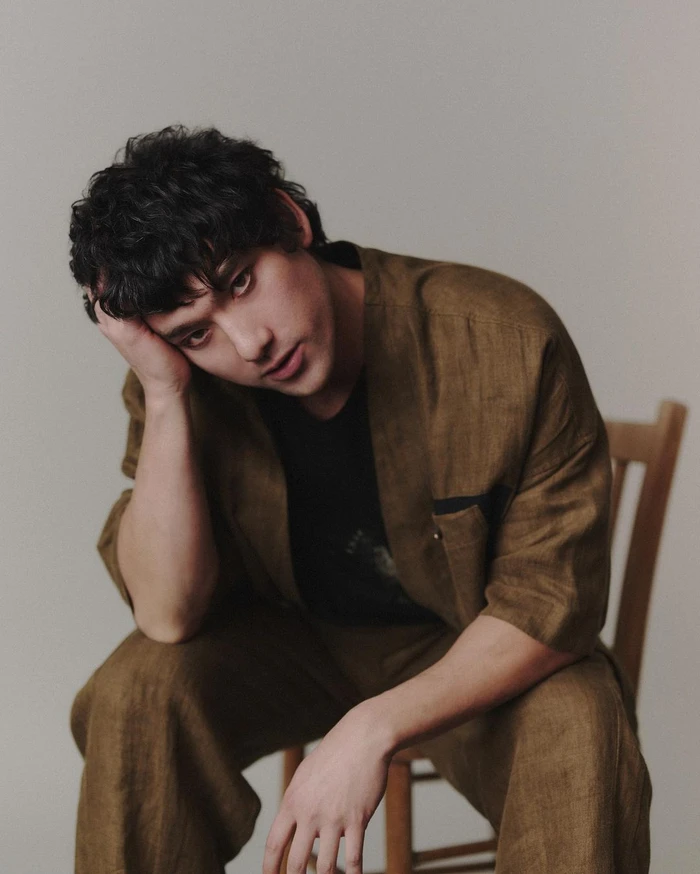 Portrait of a man with dark curly hair wearing a brown linen suit jacket and trousers, seated on a wooden chair against a plain light wall.