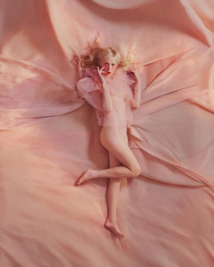 Blonde woman reclining on ruffled peach fabric wearing a sheer ruffled pink top and red lipstick