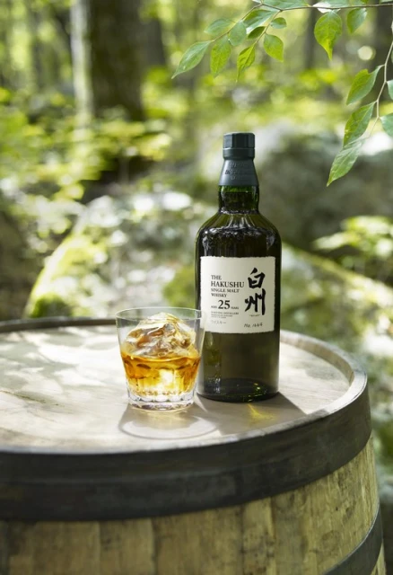Hakushu 25 Year Old single malt whisky bottle and a glass with ice on a barrel top in a forest setting.