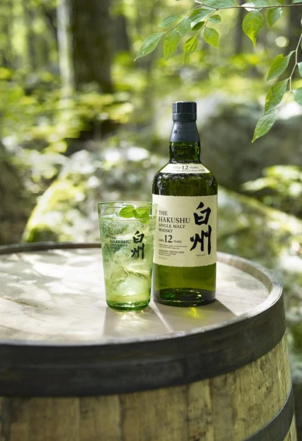 Bottle and glass of Hakushu 12 year single malt whisky with ice and mint on a wooden barrel in a sunlit forest setting.