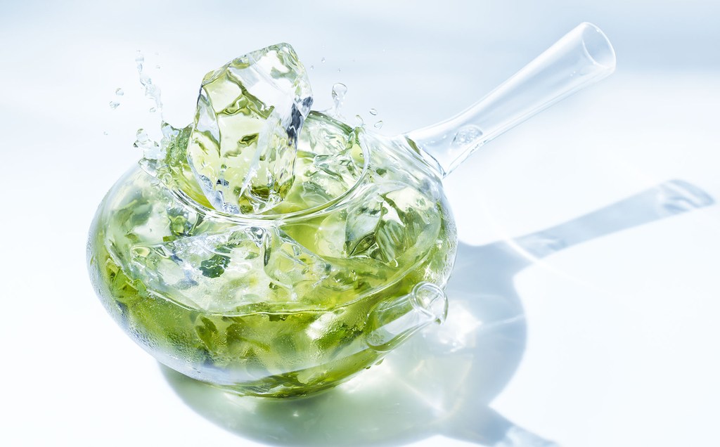 Clear teapot with green liquid and ice cubes creating splash effect, highlighted by a bright white backdrop and subtle shadow.