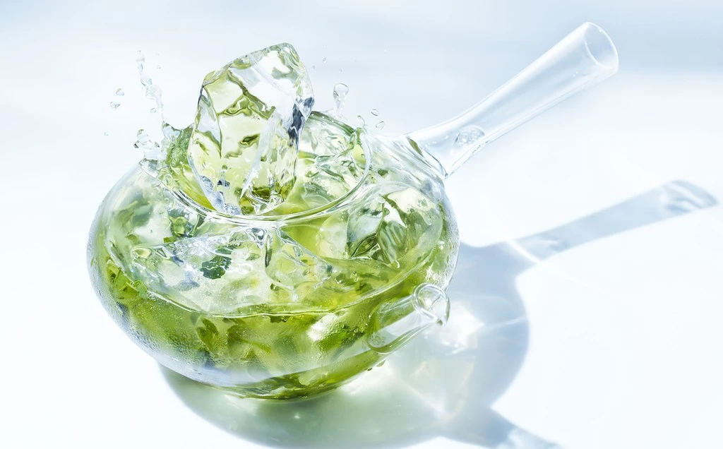 Clear teapot with green liquid and ice cubes creating splash effect, highlighted by a bright white backdrop and subtle shadow.