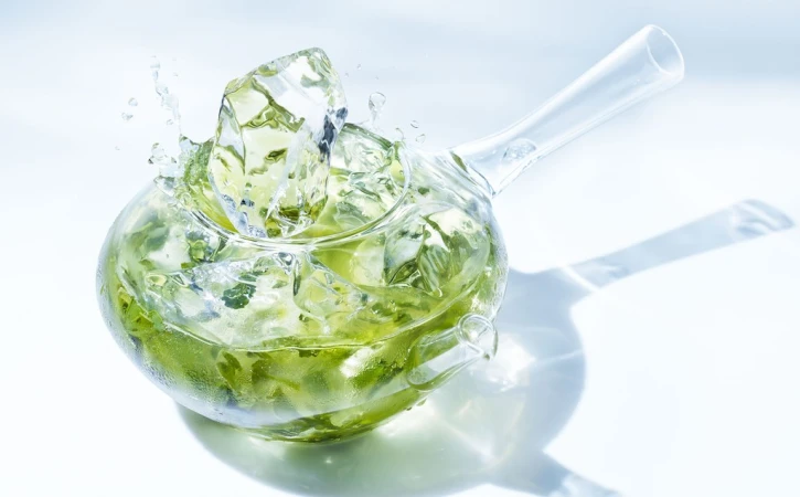 Clear teapot with green liquid and ice cubes creating splash effect, highlighted by a bright white backdrop and subtle shadow.