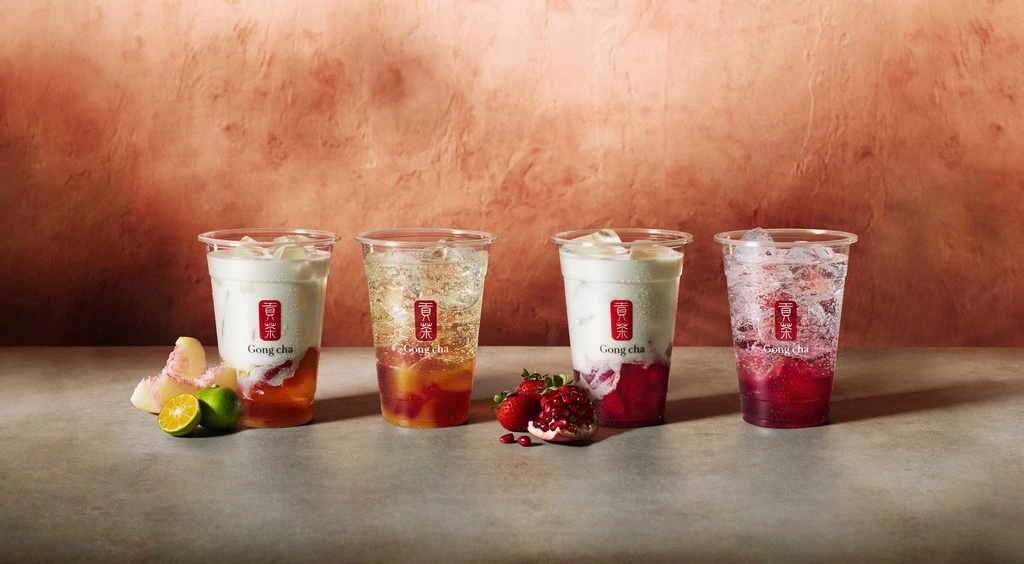 Four clear cups of colorful fruit drinks with ice and Gong Cha logos on a beige surface and reddish-brown textured backdrop.