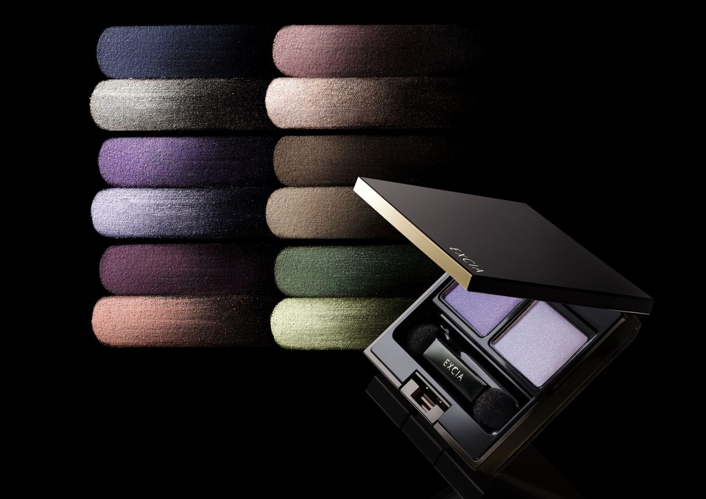 Open eyeshadow palette with makeup swatches in shades of purple, green, and brown. Dark background with dramatic lighting.