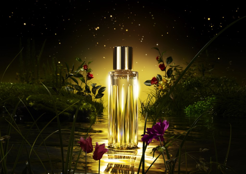 Product shot of gold-toned perfume bottle with text label in lush green and floral pond environment against a bokeh-lit dark background.