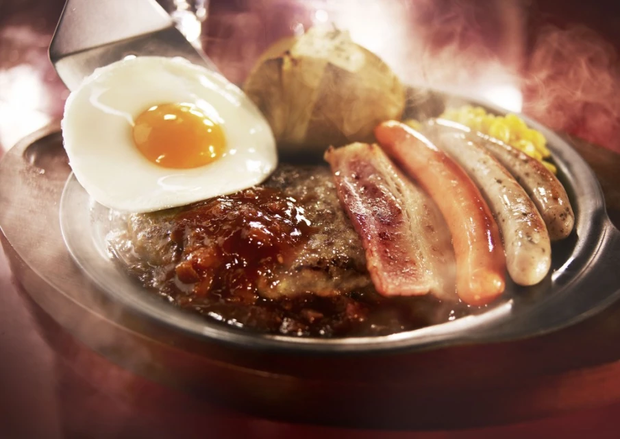 Plated meal features a juicy hamburger patty topped with a sunny-side-up egg, accompanied by bacon, sausages, corn, and a baked potato.