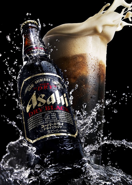 Asahi Dry Black beer bottle and a glass of beer with foam, photographed in a studio against a black background with splashing water.