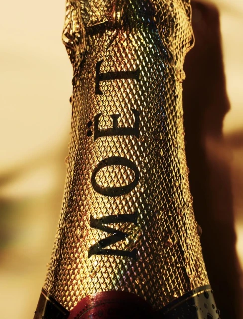 Close-up of a gold-foil-wrapped champagne bottle featuring the Moët & Chandon logo, highlighted by studio lighting and water droplets.