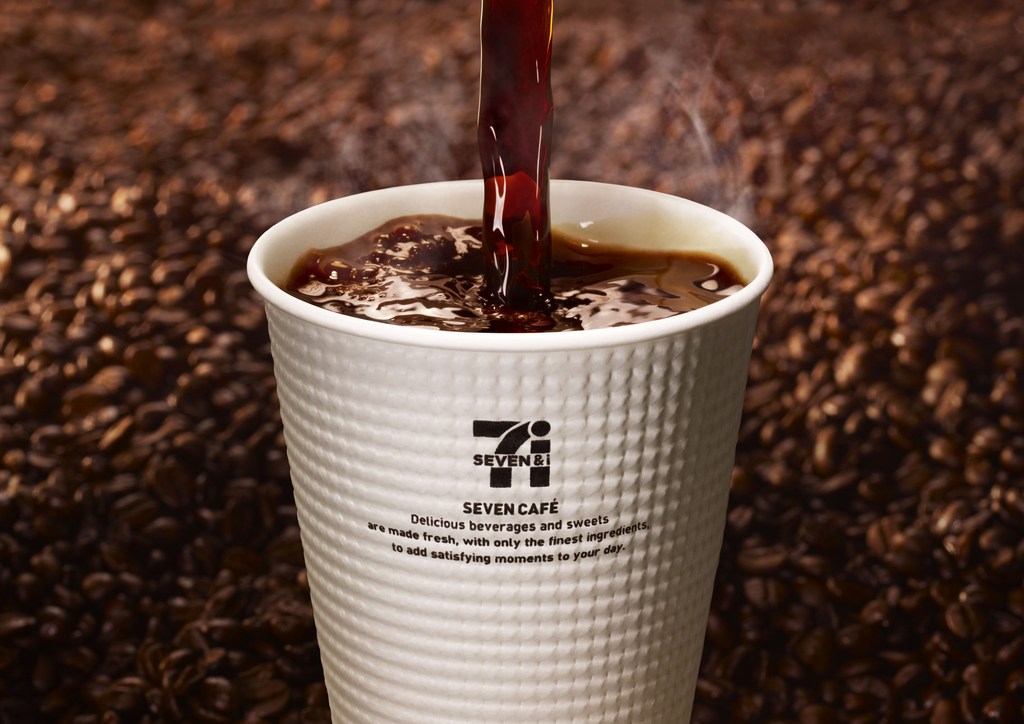 Coffee is being poured into a white paper cup with the Seven Cafe logo, set against a background of coffee beans and soft lighting.