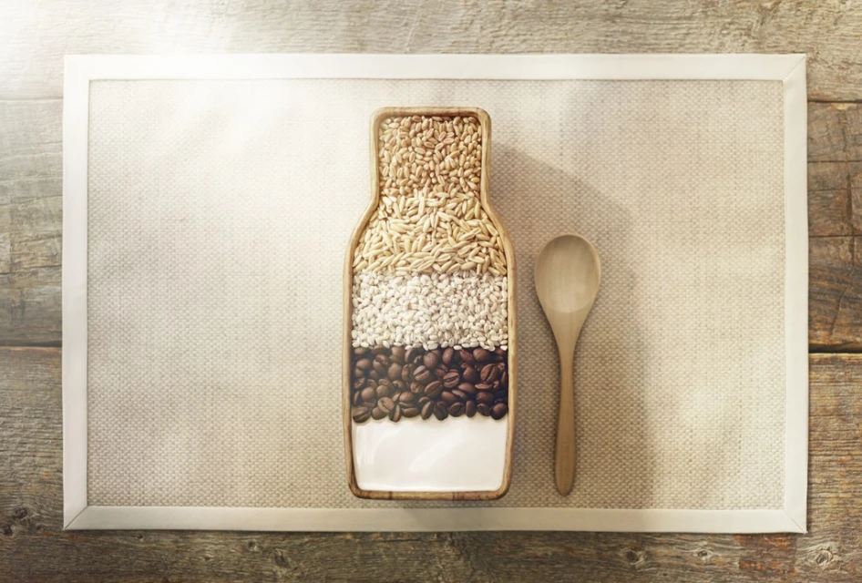 Top-down shot of grains and coffee beans in a bottle-shaped dish next to a wooden spoon, all resting on a textured placemat.