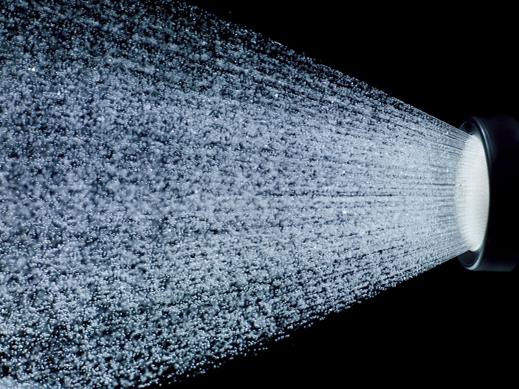 Close-up of water spraying from a dark-colored shower head against a black background, creating a high-contrast abstract image.