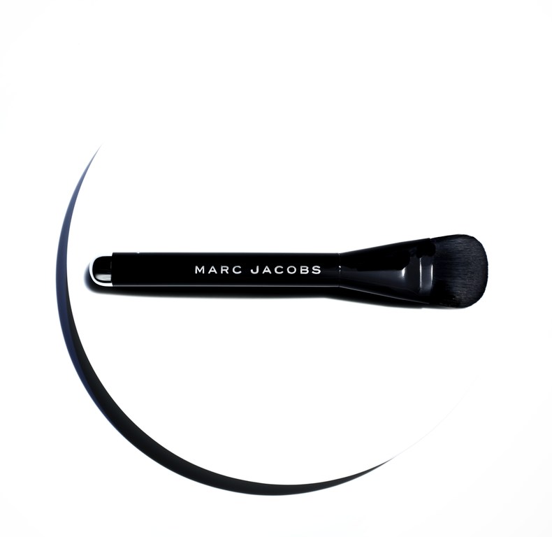 Close-up shot of a black Marc Jacobs makeup brush on a white surface with dark curved lines suggesting shadows, highlighting its design.
