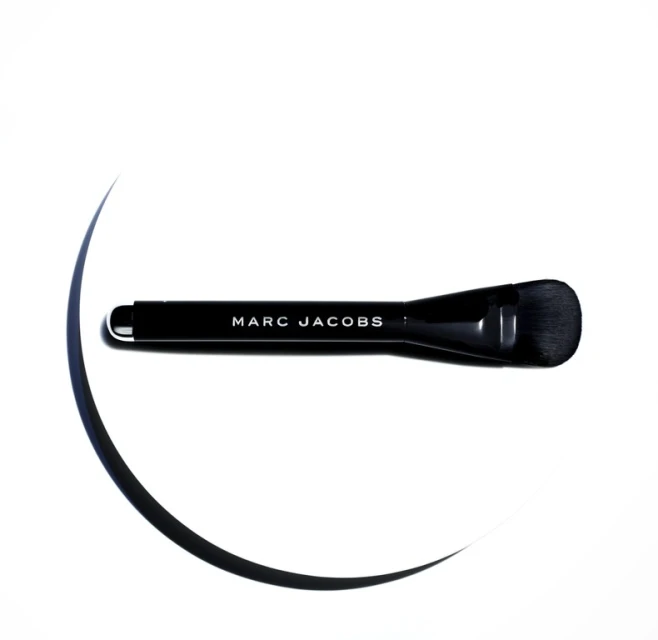 Close-up shot of a black Marc Jacobs makeup brush on a white surface with dark curved lines suggesting shadows, highlighting its design.