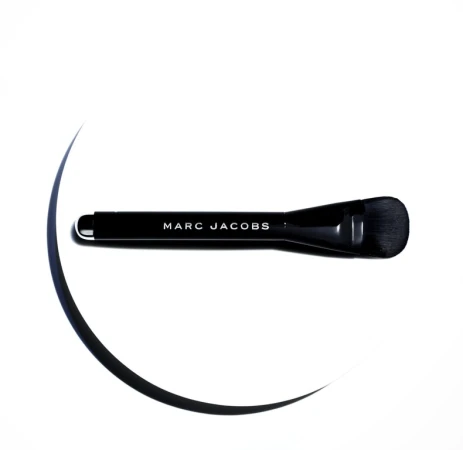 Close-up shot of a black Marc Jacobs makeup brush on a white surface with dark curved lines suggesting shadows, highlighting its design.