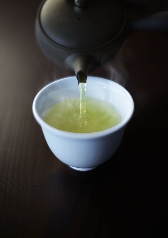Pouring hot green tea from a black teapot into a white teacup on a dark wooden surface; steam rising.