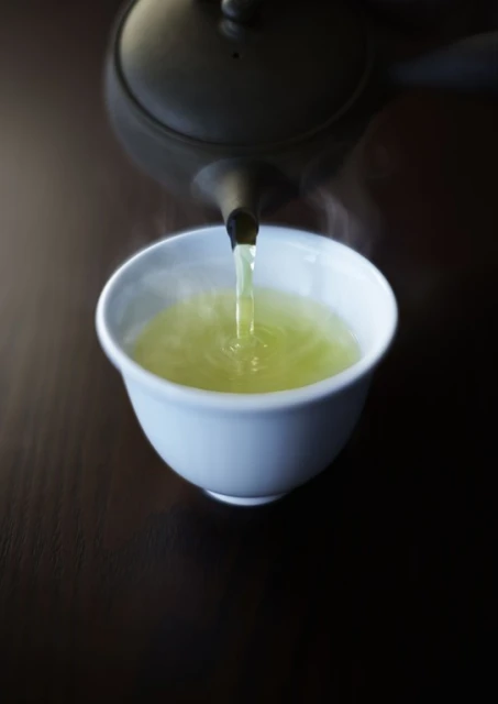 Pouring hot green tea from a black teapot into a white teacup on a dark wooden surface; steam rising.