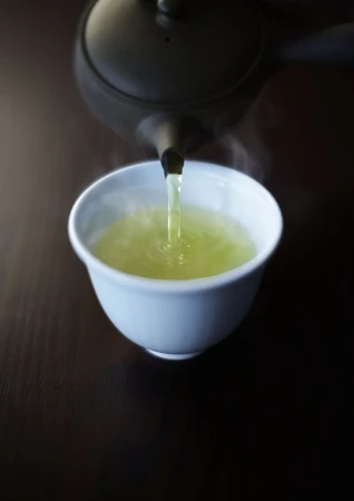 Pouring hot green tea from a black teapot into a white teacup on a dark wooden surface; steam rising.