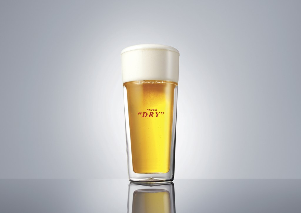 A clear pint glass filled with light beer and topped with white foam against a gradient gray background. Studio lighting.