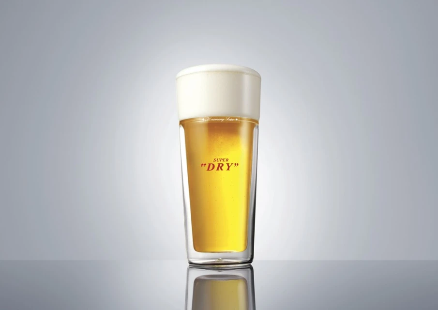 A clear pint glass filled with light beer and topped with white foam against a gradient gray background. Studio lighting.