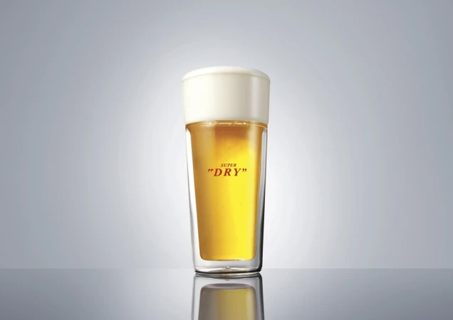 A clear pint glass filled with light beer and topped with white foam against a gradient gray background. Studio lighting.