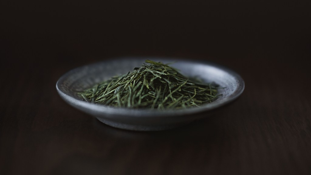 Elevated, close-up studio shot of a black ceramic dish filled with a pile of dry green tea leaves, set against a dark brown backdrop.