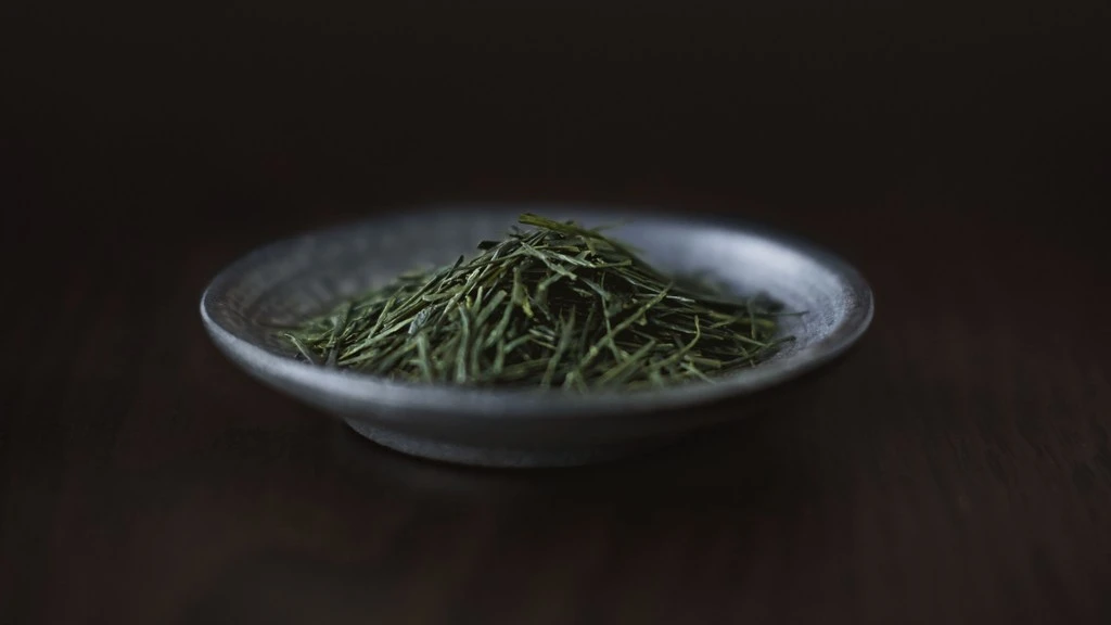 Elevated, close-up studio shot of a black ceramic dish filled with a pile of dry green tea leaves, set against a dark brown backdrop.