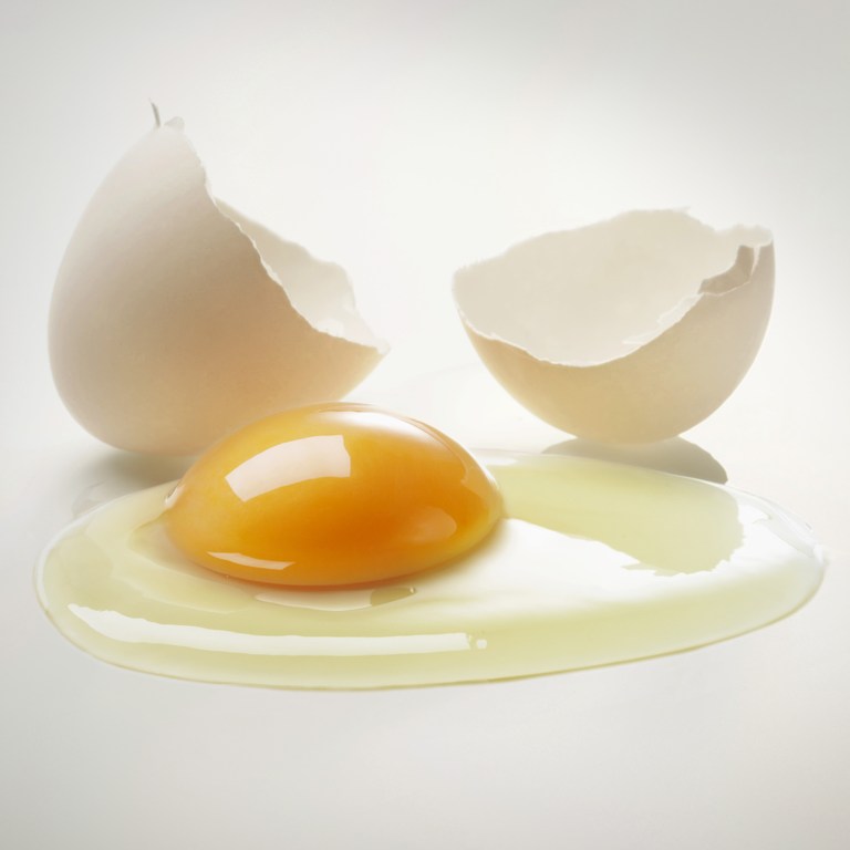 Close-up food shot of an egg with a cracked shell and a bright yellow yolk sitting in the egg white on a light background.