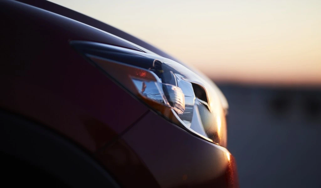 Close-up shot of a red car's headlight showcasing sleek automotive design against a blurred warm-toned sunset backdrop.
