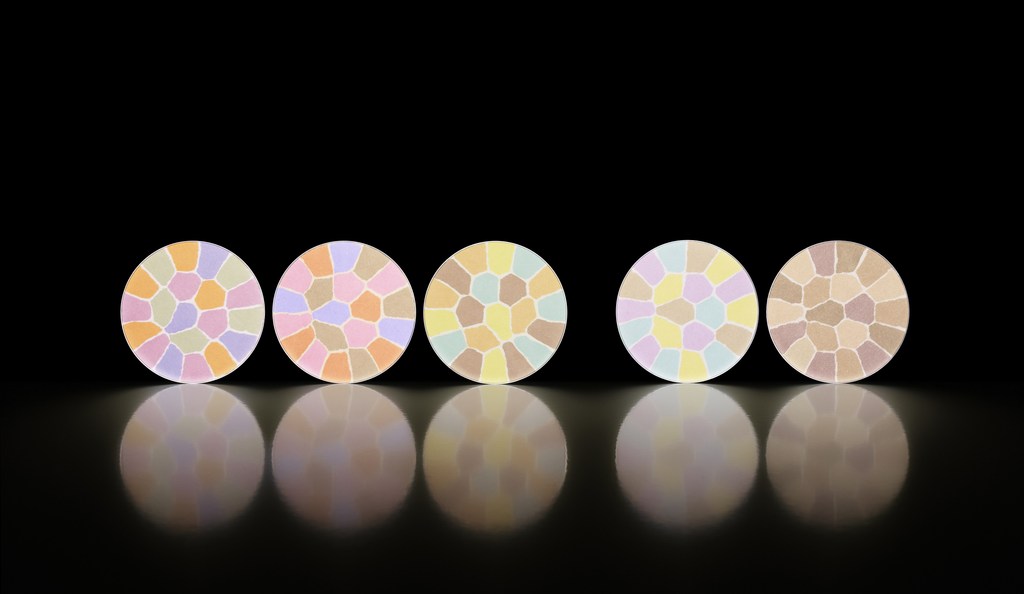 Five round cosmetic compacts with colorful mosaic patterns are arranged on a reflective surface against a stark black background.